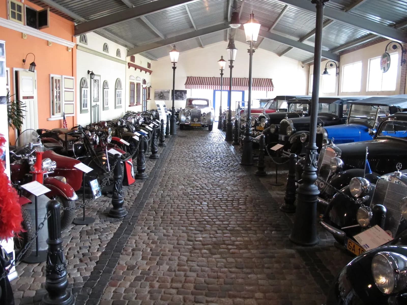 A long, covered hallway with a cobblestone floor, lined on both sides with vintage motorcycles and classic cars.