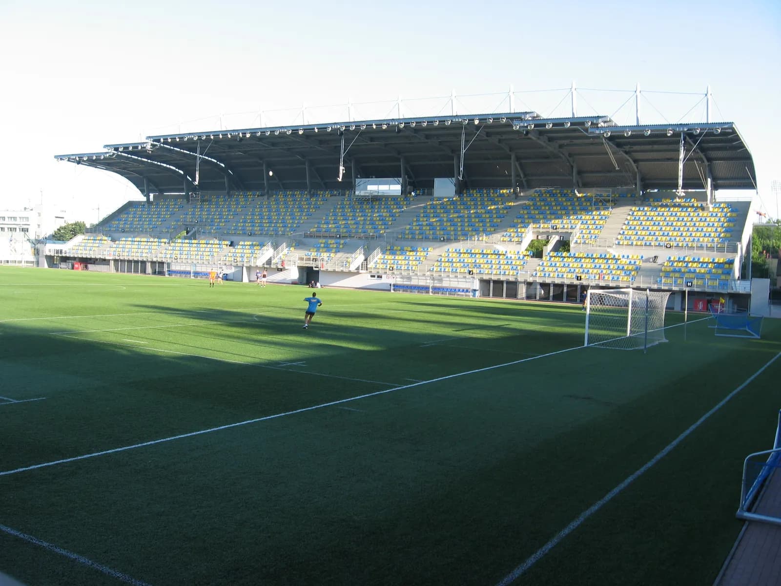 A wide view of a soccer stadium with green artificial turf, empty blue and yellow seats under a large metal roof, and a single player in a blue shirt running on the field.